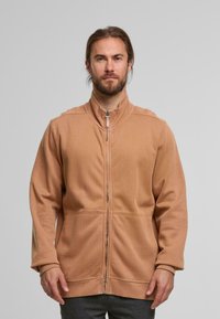 Beige Zip-Jacke mit hohem Kragen, weichem Material, langen Ärmeln und gerippten Bündchen. Verfügt über einen zentralen Reißverschluss und ein minimalistisches Design.