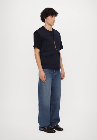 Gilet navy à manches courtes, fermeture éclair, et plusieurs poches avant. Associé à un jean bleu ample et des chaussures sombres.