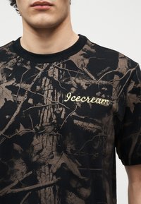 Νεαρό άτομο που φοράει μαύρο και καφέ μπλουζάκι με σχέδιο παραλλαγής και το κείμενο «Icecream» τυπωμένο στο στήθος.