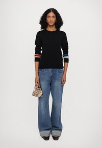 Svart genser med flerfargede stripete mansjetter, kombinert med løse blå jeans og en mønstrete håndveske, som viser uformelle, lagdelte teksturer.