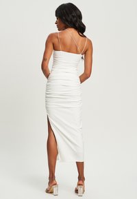 White, fitted dress with spaghetti straps, ruching details, and a thigh-high slit. Smooth texture and mid-length silhouette. Clear block-heeled sandals.