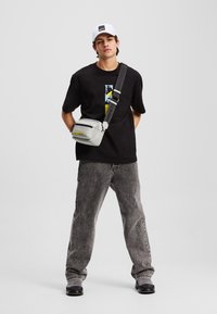 Grå crossover-veske med svarte detaljer, rektangulær form; kombinert med en svart t-skjorte og grå jeans. Modellen har på seg en hvit caps.
