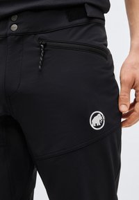 Nærbilde av svarte bukser med glidelåslomme, knappelukking og hvitt mammutlogo på låret, hånd som hviler ved siden av lommen.