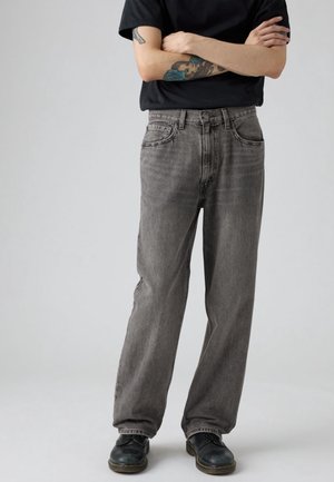 Personne portant un jean gris délavé, des chaussures noires et une chemise noire à manches courtes avec les bras tatoués croisés.