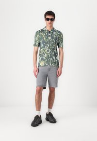 Kortermet skjorte med grønt og gult abstrakt mønster, kombinert med grå denimshorts og svarte joggesko. Solbriller fullfører stilen.