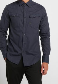 Knapp-up skjorta i mörk marinblått tyg med långa ärmar, två bröstfickor och tryckknappar. Har en krage och subtila sömdetaljer.