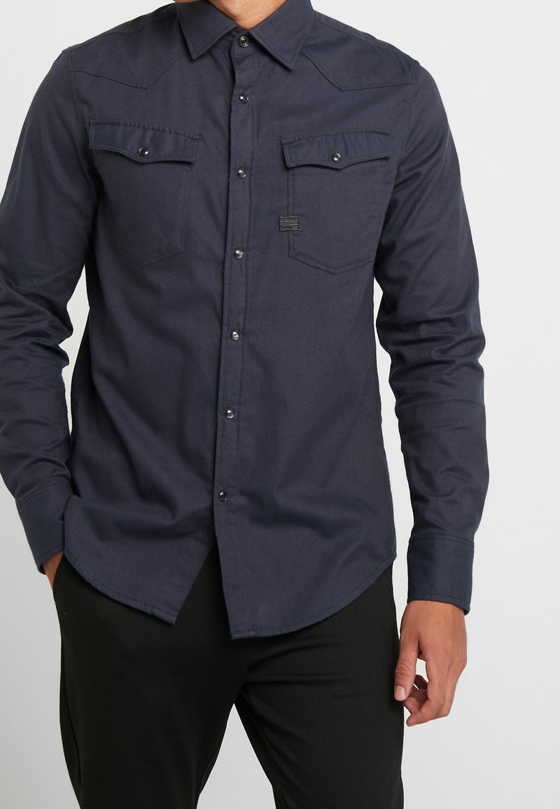 Knapp-up skjorta i mörk marinblått tyg med långa ärmar, två bröstfickor och tryckknappar. Har en krage och subtila sömdetaljer.