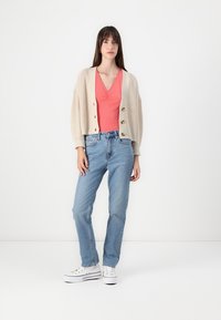 Kremfarget strikkegenser med drop shoulders og knapper, sammen med en korallfarget V-hals topp og lyseblå denimjeans. Hvite sneakers fullfører antrekket.