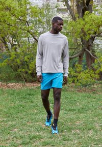 Maglietta grigia a maniche lunghe, pantaloni sportivi blu, calze nere e scarpe da ginnastica blu. In piedi sull'erba con alberi verdi sullo sfondo.