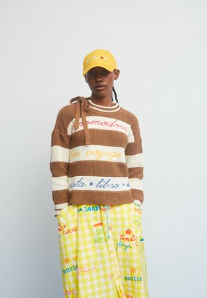 Strikket brun og cremefarvet stribet sweater med tekstdetaljer, kombineret med et gulternede skørt med farverige tekster og grafik, samt en gul kasket.