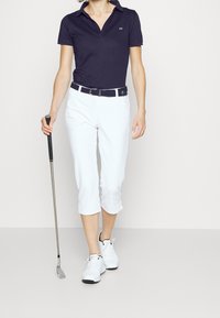 Tmavě modré polo tričko s límečkem a logem, v kombinaci s bílými zkrácenými kalhotami a bílými golfovými botami. Drží golfovou hůl.