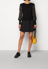 Maglia nera a maniche lunghe con accenti di punti dorati sulle maniche, abbinata a una gonna skater nera. Gli accessori includono una borsa gialla e scarpe nere.