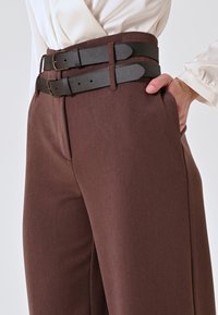 Bruine hoge taillebroek met twee overlappende zwarte leren riemen met rechthoekige gespen, zijzakken en een getailleerde pasvorm.