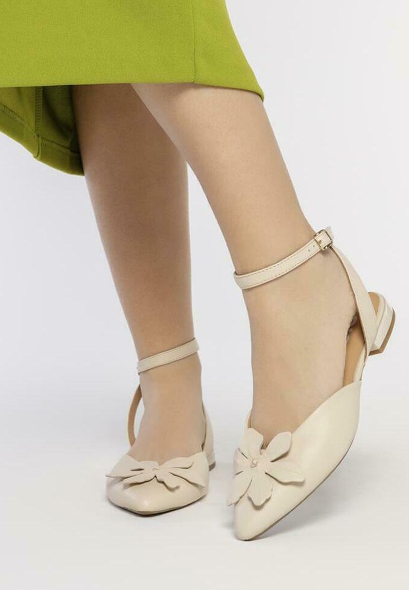 Bailarinas de cuero beige con puntera afilada, con un acento decorativo de flor y una delgada tira en el tobillo, sobre un fondo claro.