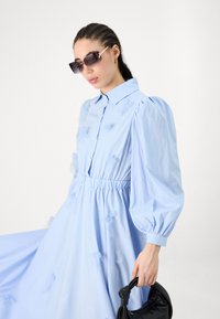 Φόρεμα σε ανοιχτό μπλε χρώμα με σχέδιο που έχει γιακά, φουσκωτά μανίκια και συγκεντρωμένη μέση. Διαθέτει διακοσμητικά από ύφασμα και λεία υφή. Μαύρη τσάντα χεριού.