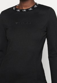 Svart långärmad klänning med en åtsittande silhuett, texturerad halsringning med kontrasterande cK-logotyp och subtil Calvin Klein-branda på framsidan.