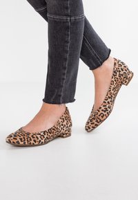 Femme portant des chaussures à talons bas en imprimé léopard et un jean noir avec des ourlets effilochés, se tenant sur un sol blanc.