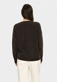 Frau mit langen dunklen Haaren, die einen dunkelbraunen Pulli und helle beigefarbene Hose trägt, von hinten vor einem schlichten Hintergrund gezeigt.