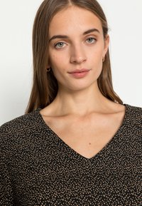 Peça de vestuário preta em formato de V com um padrão texturizado em manchas bege, apresentando um corte relaxado e um tecido suave. Brincos de argola dourados complementam o visual.