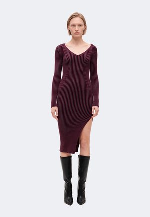 Femme portant une robe midi à manches longues rayée violet et noir avec une fente haute sur la cuisse et des bottes noires montantes, debout face à nous.