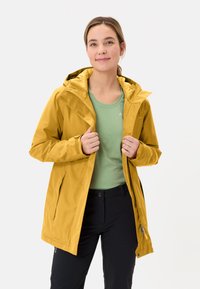 Giacca impermeabile gialla con cappuccio, tasche laterali con zip e polsini regolabili, indossata sopra una camicia verde chiaro e pantaloni neri.