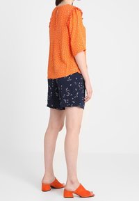 Lys orange bluse med korte ærmer, tekstureret stof, parret med marineblå shorts med blomstermønster. Orange blokhælede sandaler.
