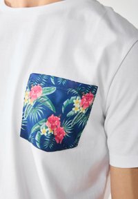 White T-shirt featuring a navy blue pocket with a tropical floral pattern, including pink and yellow flowers and green leaves.