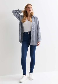 Camisa de botones a rayas en azul y blanco, llevada sobre una camiseta blanca sin mangas, combinada con jeans oscuros de tiro alto y zapatillas blancas.