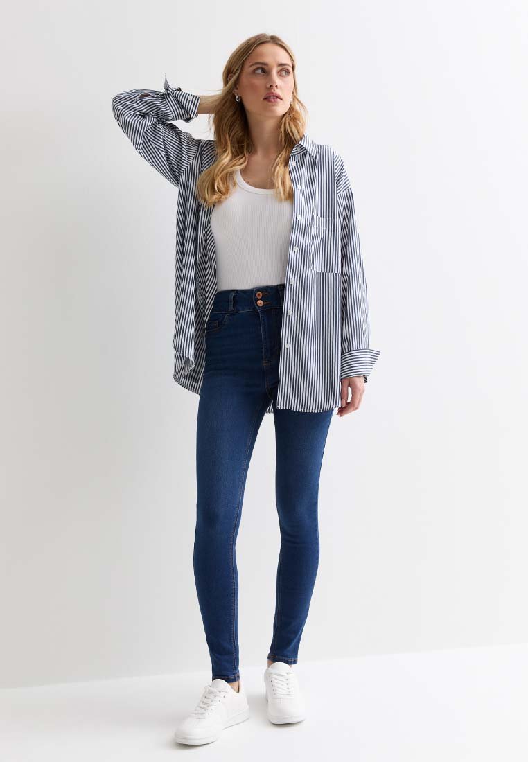 Camisa de botones a rayas en azul y blanco, llevada sobre una camiseta blanca sin mangas, combinada con jeans oscuros de tiro alto y zapatillas blancas.