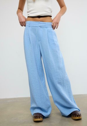 Osoba nosiaca voľné, svetlomodré textúrované nohavice s vysokým pásom a širokými nohavicami, doplnené o hnedé dreváky na platforme, upravujúca si pás.