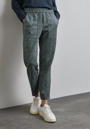 Person trägt gemusterte dunkle Hosen mit elastischem Bund und weiße Turnschuhe, steht vor einem einfarbigen hellen Hintergrund mit den Händen in den Taschen.