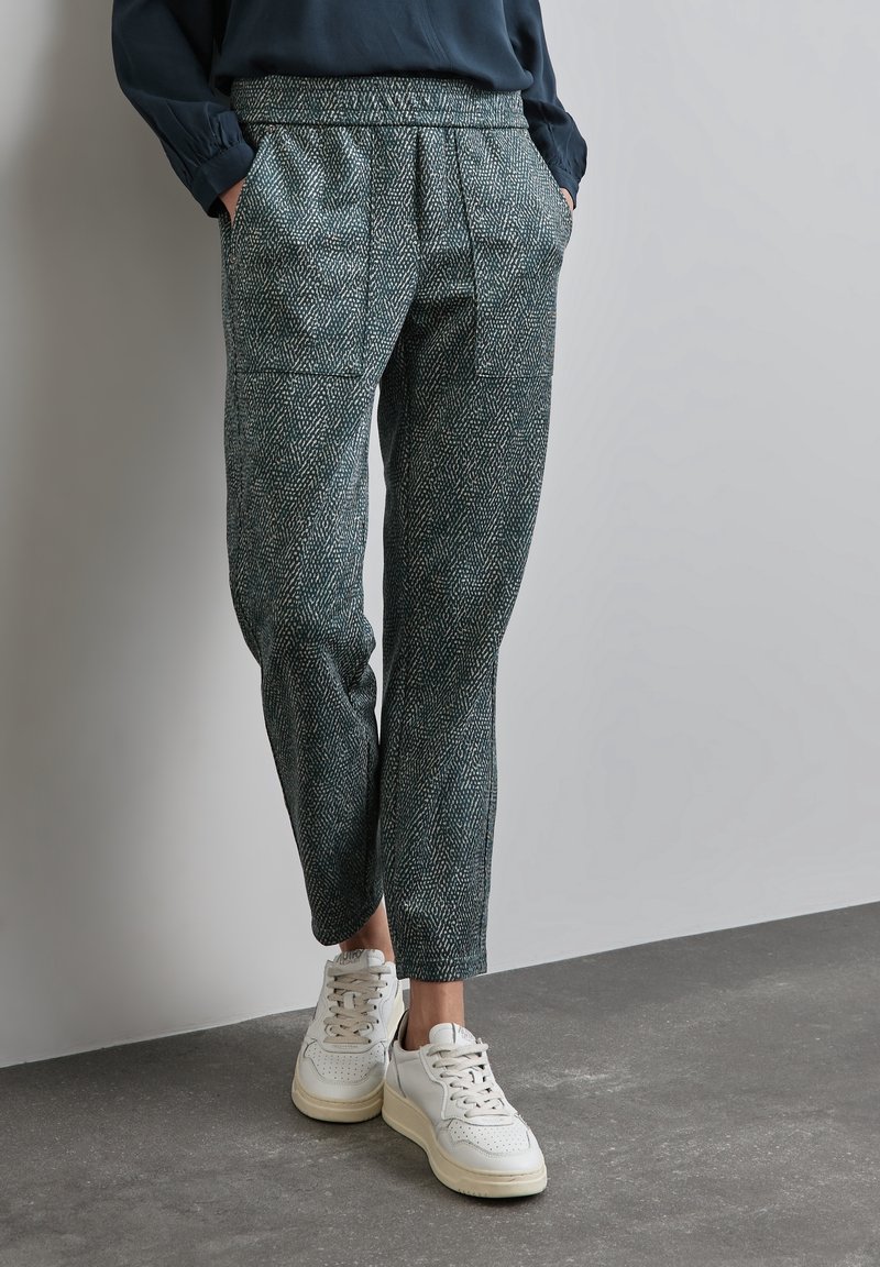 Personne portant un pantalon foncé à motif avec une taille élastique et des baskets blanches, debout devant un fond clair uni, les mains dans les poches.