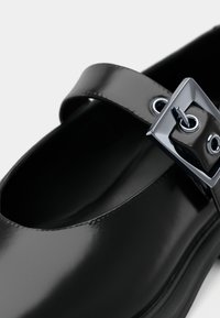 Black patent leather shoe with rounded toe, featuring a wide strap and a rectangular metallic buckle with eyelets. Smooth, shiny texture.
