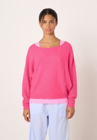 Pull surdimensionné rose vif avec un large décolleté et des manches longues, fabriqué à partir d'un matériau doux et texturé ; porté par-dessus un haut rose clair.