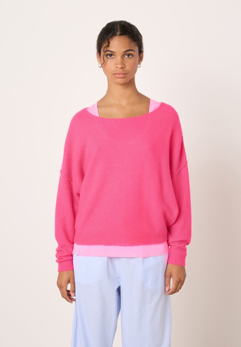 Pull surdimensionné rose vif avec un large décolleté et des manches longues, fabriqué à partir d'un matériau doux et texturé ; porté par-dessus un haut rose clair.