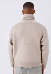 Hombre de espaldas vestido con un suéter de cuello alto beige y jeans de color azul claro con bolsillos traseros, contra un fondo liso.
