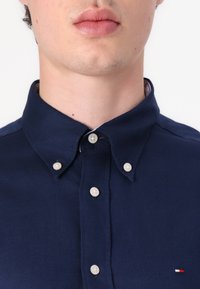 Cămașă albastră cu nasturi, cu guler, având nasturi albi și un mic logo în roșu, alb și albastru pe piept. Material texturat.