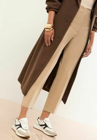 Pantaloni beige a vita alta con accenti marroni ai lati abbinati a sneakers bianche e beige. Un soprabito marrone è appoggiato accanto. Bracciale dorato al polso.