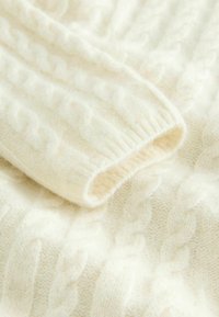 Pull en tricot de couleur crème avec un motif de torsades texturé et des poignets côtelés. Matière douce, semblable à de la laine, avec une apparence confortable.