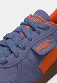 Μπλε σουέντ αθλητικό παπούτσι Puma Palermo με πορτοκαλί λογότυπο, λεπτομέρειες και κορδόνια δεμένα σε φιόγκο.