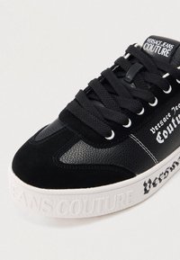 Tênis preto com atacadores e sola branca, com o texto "Versace Jeans Couture" na lateral, na língua e em relevo na sola.