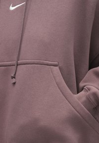 Κοντινό πλάνο χεριού στην τσέπη παλτό φούτερ Nike σε μωβ χρώμα με ορατό κορδόνι και λευκό λογότυπο Nike στη περιοχή του στήθους.