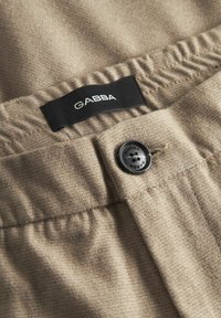 Beige szövet, textúrált felülettel, fekete gombbal és egy "GABBA" felirattal ellátott címkével. A derékpánt gyűrt kialakítású.