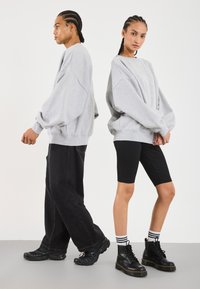 Felpe grigie oversize con spalle drop, abbinate a pantaloni corti neri e stivali neri chunky. Il modello posa in un ambiente minimalista.