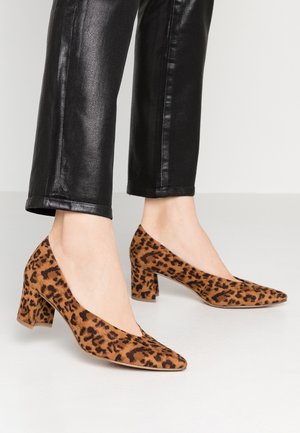 Czółenka na wysokim obcasie w panterkę z ostrym noskiem i teksturowanym zamszem, spoczywające na masywnym obcasie. W zestawie z błyszczącymi czarnymi spodniami.