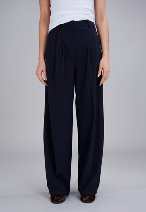 Vrouw draagt zwart hoge taille wijde broek, wit mouwloos topje en schoenen met luipaardprint, staand tegen een effen achtergrond.