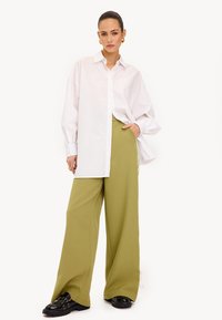 Witte oversized shirt, lichtgroene wijde broek, en zwarte schoenen met metalen accenten. Zachte stoffen, relaxte pasvorm en lange mouwen.