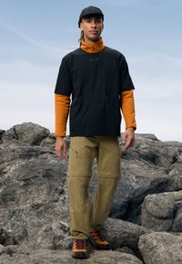 Črna kratka majica s kratkimi rokavi, položena čez oranžno majico z dolgimi rokavi, bež cargo hlače in oranžni pohodniški čevlji, na skalnatem terenu.