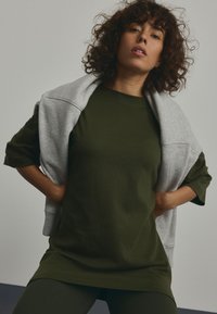 Olive green oversized t-shirt with short sleeves, paired with a light gray sweatshirt draped over the shoulders. Smooth fabric texture.