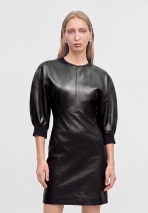 Femme aux longs cheveux blonds portant une robe en cuir noire ajustée avec des manches trois-quarts bouffantes, sur un fond clair uni.